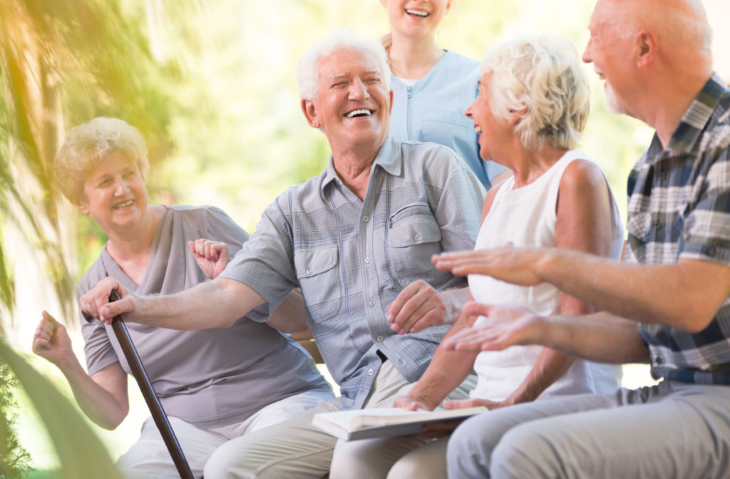 A group of seniors are sitting together on a bench, smiling and looking directly at the camera, with a nurse standing behind them