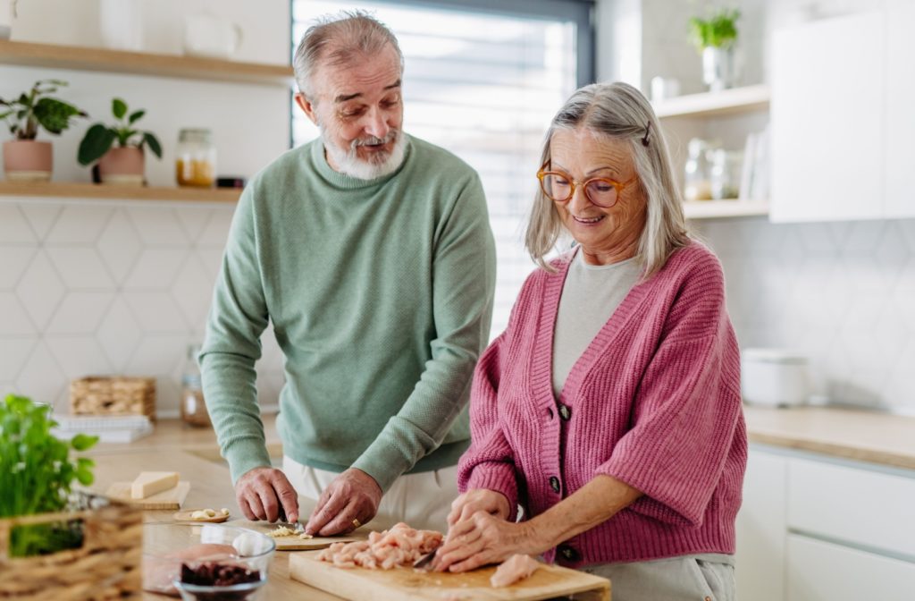 An older married couple in a sunlit kitchen cutting up a chicken to make a senior-friendly keto meal.